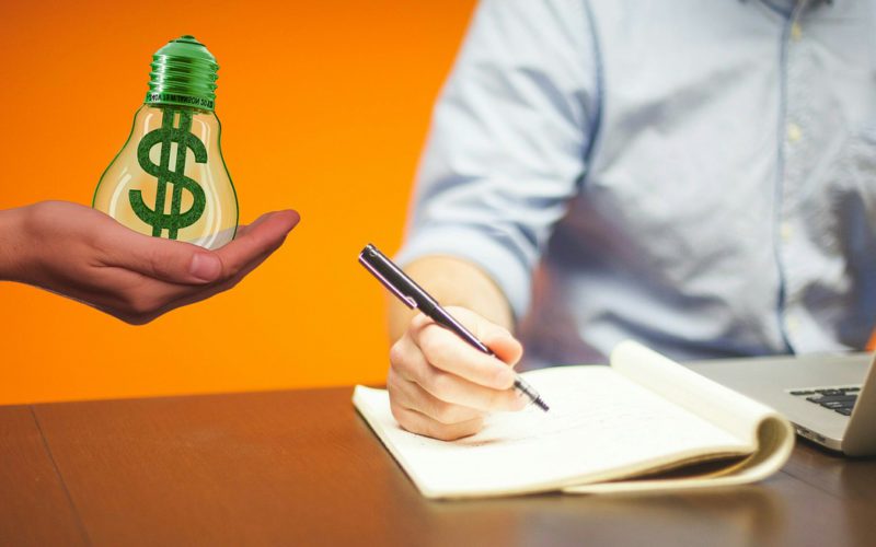 Person writing in notebook is being handed a lightbulb with a dollar sign in it.